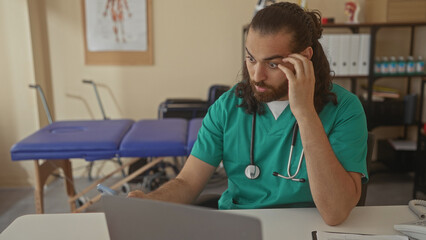 Man doctor with stethoscope rubbing eyes and massaging temples at clinic desk while reviewing documents and laptop; tired stress burnout.