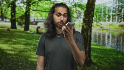 Young man with long hair and beard speaking into smartphone held in right hand in forest by a riverside pond; surprise voice assistant.