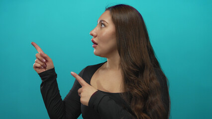 Obraz premium Young hispanic woman with long brown hair pointing finger to object in studio against a turquoise wall with slightly open mouth; surprise curiosity discovery wonder.