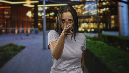 Fototapeta premium Woman pointing finger at camera and holding finger to lips for silence on street; secrecy seriousness.