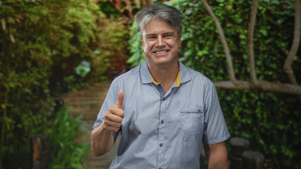 Man giving thumbs up with visible hand in forest setting wearing short sleeve shirt, smiling;...