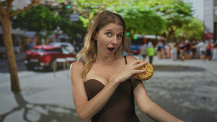 Fototapeta premium Woman holding a sponge in one hand and a loofah in the other, arms raised on a busy city street in a brown swimsuit showing cleavage; indecision.