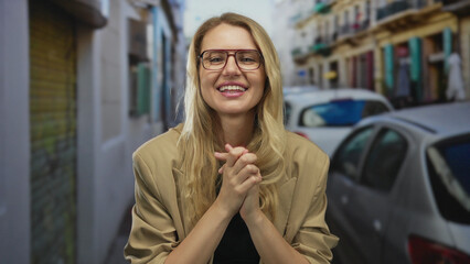Smiling blonde woman with glasses in casual attire stands confidently on a bustling city street...