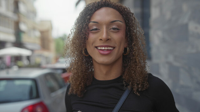 Transgender woman with curly hair points finger upward on city street lined with parked cars and stone facade; inspiration.