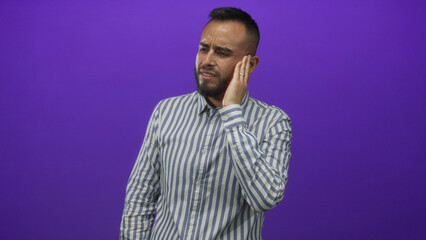 Young hispanic man in a striped shirt holding hand to ear and wincing in a purple studio with toothache; pain.