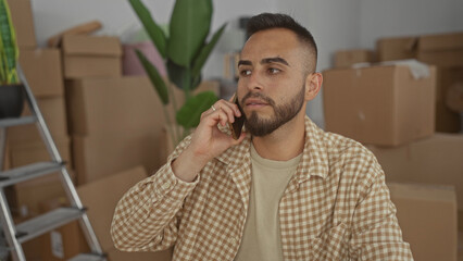 Man holds phone to ear and gestures with free hand amid packed moving boxes, ladder and houseplant in a building room; moving day stress.