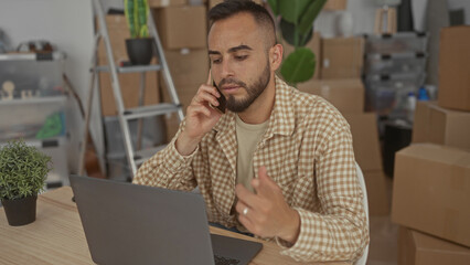 Man talking on phone at home at a table with laptop and hand on keyboard, packed boxes and ladder visible; moving productivity.