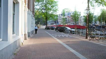Blurred cityscape in amsterdam featuring a lone man walking along a canal-side path lined with bicycles and traditional dutch architecture in defocused background.