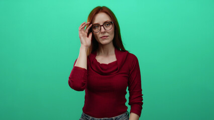 Woman with red hair and glasses in red sweater against green wall, expression thoughtful and confident, isolated setting emphasizing modern and stylish appearance