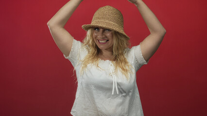 Woman wearing straw hat and white blouse raising arms above head and smiling in studio with red...