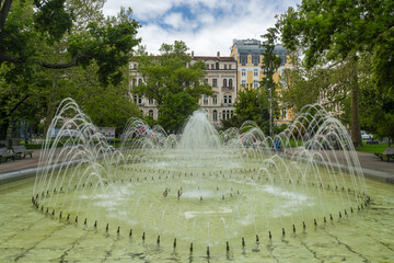 View of a symmetrical fountain spraying water in a park with trees and buildings in the background, under a blue sky, Sofia, Sofia City Province, Bulgaria.