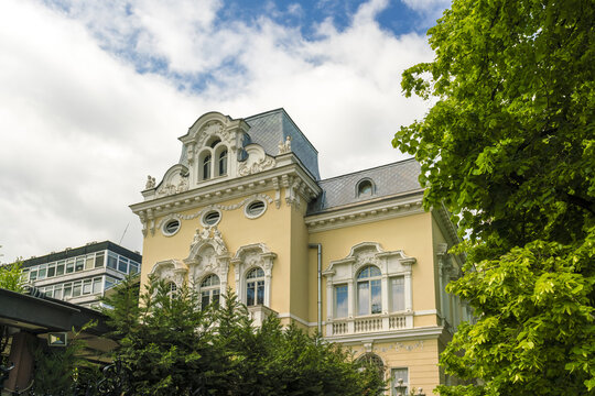 View of an ornate yellow building with white trim, detailed architectural features, and green trees against a blue sky with clouds, Sofia, Sofia City Province, Bulgaria.