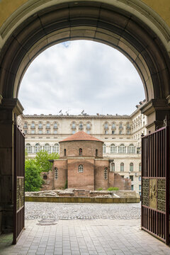 View of an ancient rotunda, framed by an arched gateway, stands in contrast to the modern building behind, a blend of history and progress, Sofia, Sofia City Province, Bulgaria.