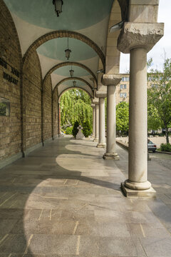 View of the colonnade's cool shadows playing across the stone floor, contrasting with the bright greenery and sky beyond the arches, Sofia, Sofia City Province, Bulgaria.