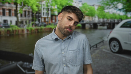 Fototapeta premium Man tilts head and pouts lips by canal on city street amid green tree canopy with buildings lining waterway; curiosity.
