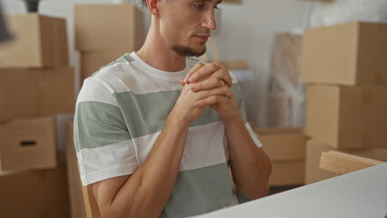 Man with hands clasped at chest, fingers interlaced amid stacked moving boxes in a residential building, pausing at a table; hope settling.