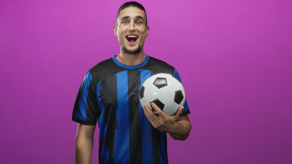 Man holding soccer ball with hand, smiling in studio wearing blue striped jersey; joyful celebration.