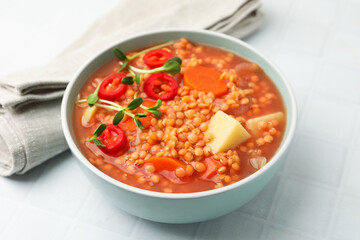Delicious lentil soup with vegetables in bowl on white tiled table, closeup