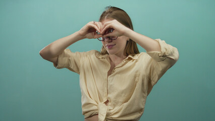 Young blonde woman makes binoculars gesture in studio with teal background; exploration curiosity...