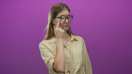 Blonde woman adjusting round glasses with slight smile by pointing finger to lens in purple studio...