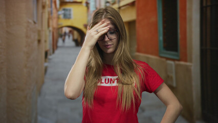 Woman covers eyes with hand in narrow street wearing volunteer tshirt with blonde hair visible; exhaustion.
