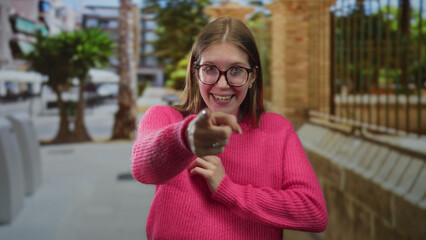Fototapeta premium Woman in pink sweater and glasses smiling and pointing finger on city street with brick building wall behind; confidence.