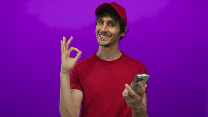 Man wearing red cap and shirt taps smartphone then shows ok sign against purple backdrop in studio; confidence approval connection communication.