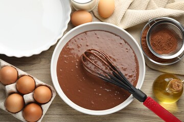 Liquid chocolate dough in bowl, whisk, oil, milk, cocoa powder and eggs on wooden table, flat lay