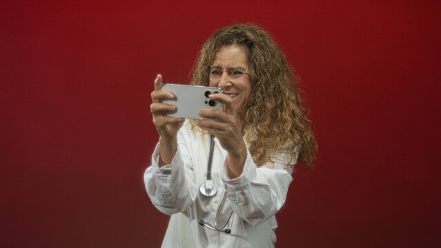 Woman doctor holds smartphone with both hands taking a selfie, stethoscope visible in white coat, studio red backdrop; telemedicine confidence. - Powered by Adobe