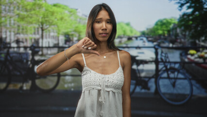 Thai woman in white dress showing thumbs down with bare thumb on a street bridge beside canal and parked bicycles; disapproval.