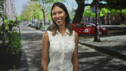 Young thai woman smiling in a white sleeveless shirt on a street with a red car, planter and tree lined pavement in an urban setting; happy casual warmth.