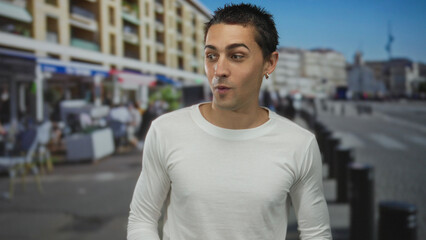 Fototapeta premium Young hispanic man in a white shirt standing on a vibrant urban street with blurred buildings and people, expressing surprise.