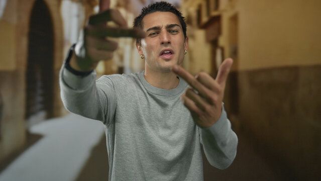Young hispanic man shows middle finger gesture in narrow cobblestone old town street; anger defiance rejection.