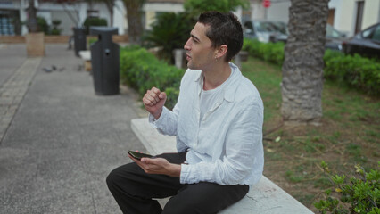 Obraz premium Young man in park wearing white shirt holds smartphone while sitting on a bench surrounded by greenery and cars parked on street.