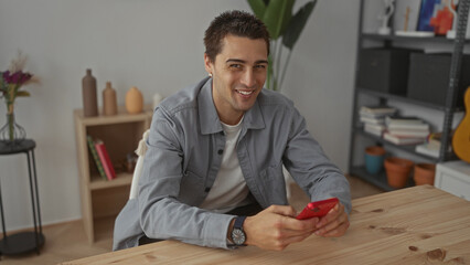 Young man using smartphone in cozy living room with guitar and bookshelves, wearing casual jacket while seated at wooden table in bright apartment interior.