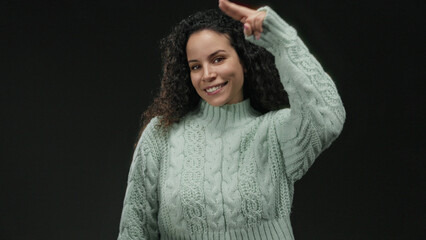 Hispanic woman in cozy sweater smiles making hand gesture against isolated black background wall in a series of frames capturing friendly expression.