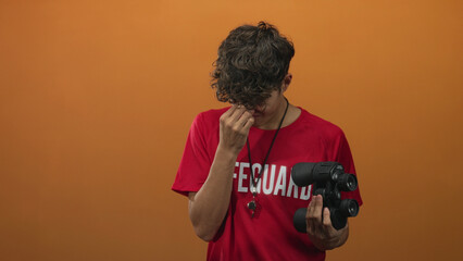 Teen lifeguard boy holding binoculars pinches nose and rubs eyes in studio with orange backdrop;...