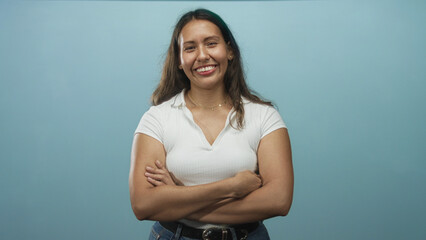 Woman with arms crossed smiling in blue studio, wearing a white top and jeans with belt, showing...
