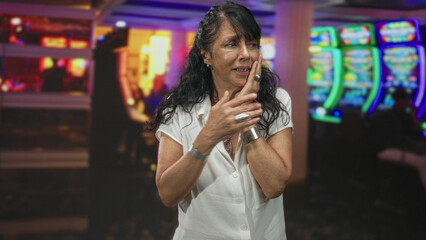 Woman senior pressing cheek with hand for toothache while pointing with other hand, wearing white blouse and rings in casino; anxiety regret loss.