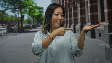 Fototapeta premium Woman points finger to open palm on city street while smiling and looking at hand; friendly invitation.