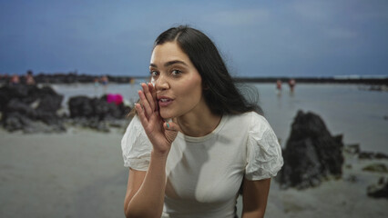 Woman whispering hand to mouth on beach with rocks and shallow water near volcanic rock pools in studio setting; playful.