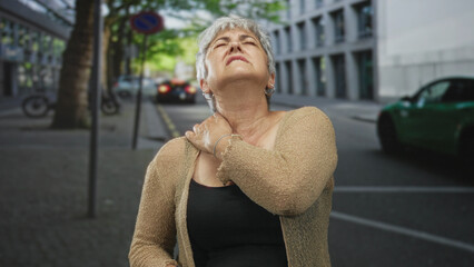 Woman holding shoulder, pressing it in visible shoulder pain while standing on a city street; discomfort aging resilience.