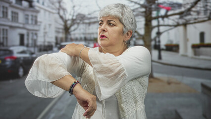 Woman with grey hair rubbing her shoulder and adjusting a sleeve while wearing bracelets on a...