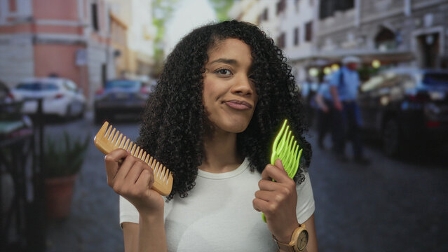 African american woman holds wooden comb and green pick as she compares hair tools on bustling street; indecision.