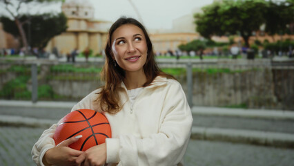 Young woman in white sweatshirt holding basketball with happy expression outdoors on a city street, surrounded by urban landscape and trees, enjoying a leisurely day.