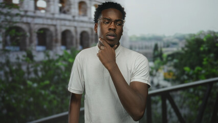 Young man finger pointing to sunlit roman coliseum building framed by green foliage and railing in...