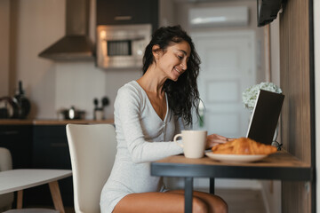 Smiling woman working on a laptop at a kitchen table with coffee and croissant nearby, enjoying morning light, comfort, focus, productivity and a cozy relaxed home atmosphere