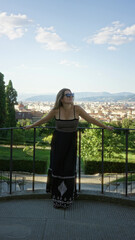 Fototapeta premium Woman standing with arms outstretched on viewpoint railing in florence wearing black sundress and sunglasses, bare feet visible; serenity.