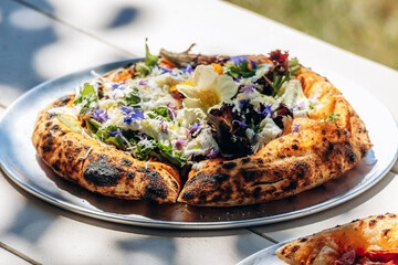 Gourmet artisan pizza with a thick, fluffy crust, decorated with edible flowers and fresh greens, served outdoors in natural sunlight.