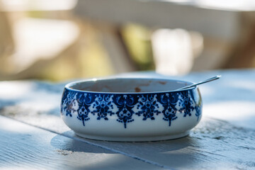A white ceramic bowl with a traditional blue ornamental pattern and a spoon inside, photographed on an outdoor wooden table in natural sunlight.